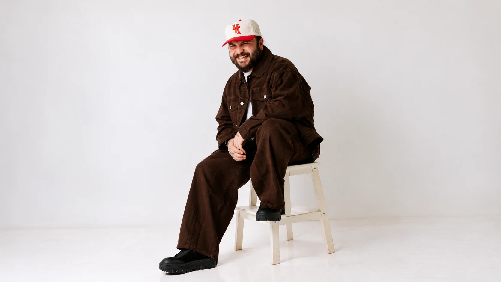 Moojo assis sur un tabouret en studio, en veste et pantalon marron
